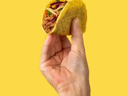 A hand holding a crispy yellow taco filled with seasoned meat and fresh vegetables, set against a bright yellow background.